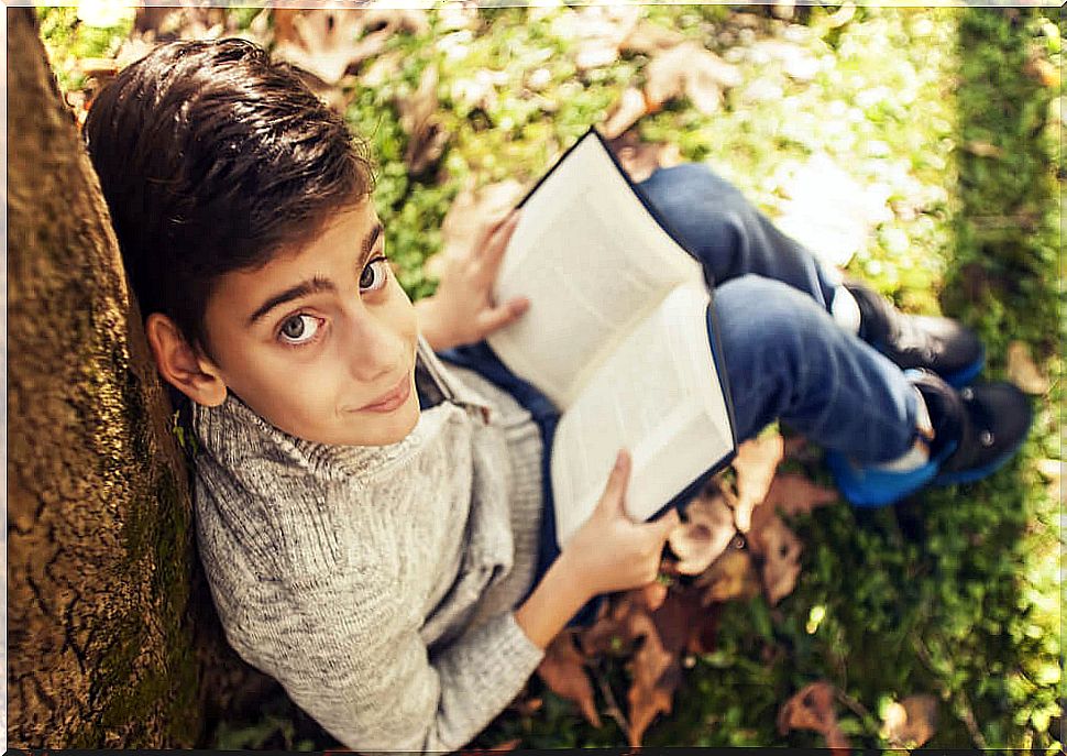 Boy reading sitting in a park with leaves on the ground.